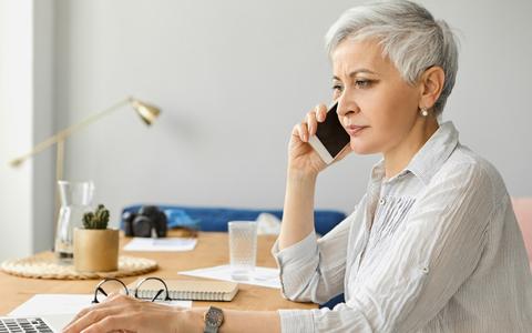 Ältere Frau mit kurzen grauen Haaren telefoniert am Schreibtisch mit ernstem Gesicht