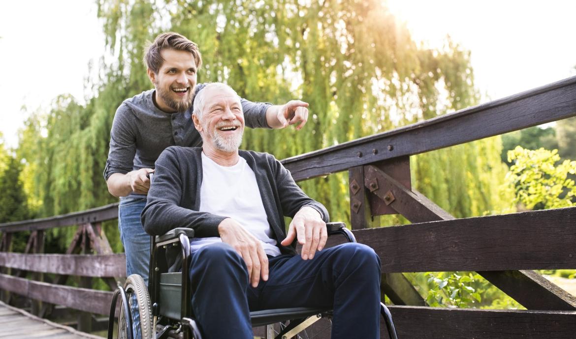 Junger Mann schiebt Rollstuhl mit älterem Herrn über eine Brücke in einem Park