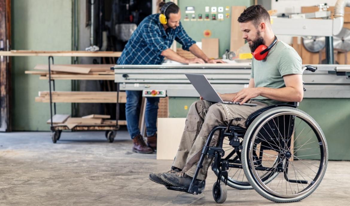Ein junger Mann sitzt in einer Werkstatt im Rollstuhl mit einem Laptop auf den Knien