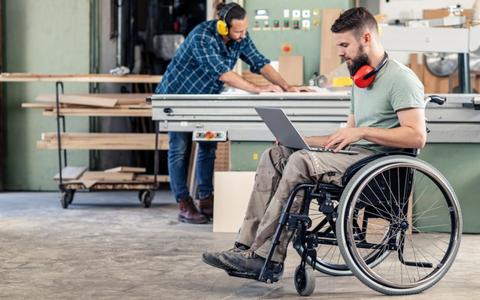 Ein junger Mann sitzt in einer Werkstatt im Rollstuhl mit einem Laptop auf den Knien